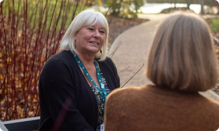 Two women sit on a bench in gardens, one listening to the other speaking and smiling at her.