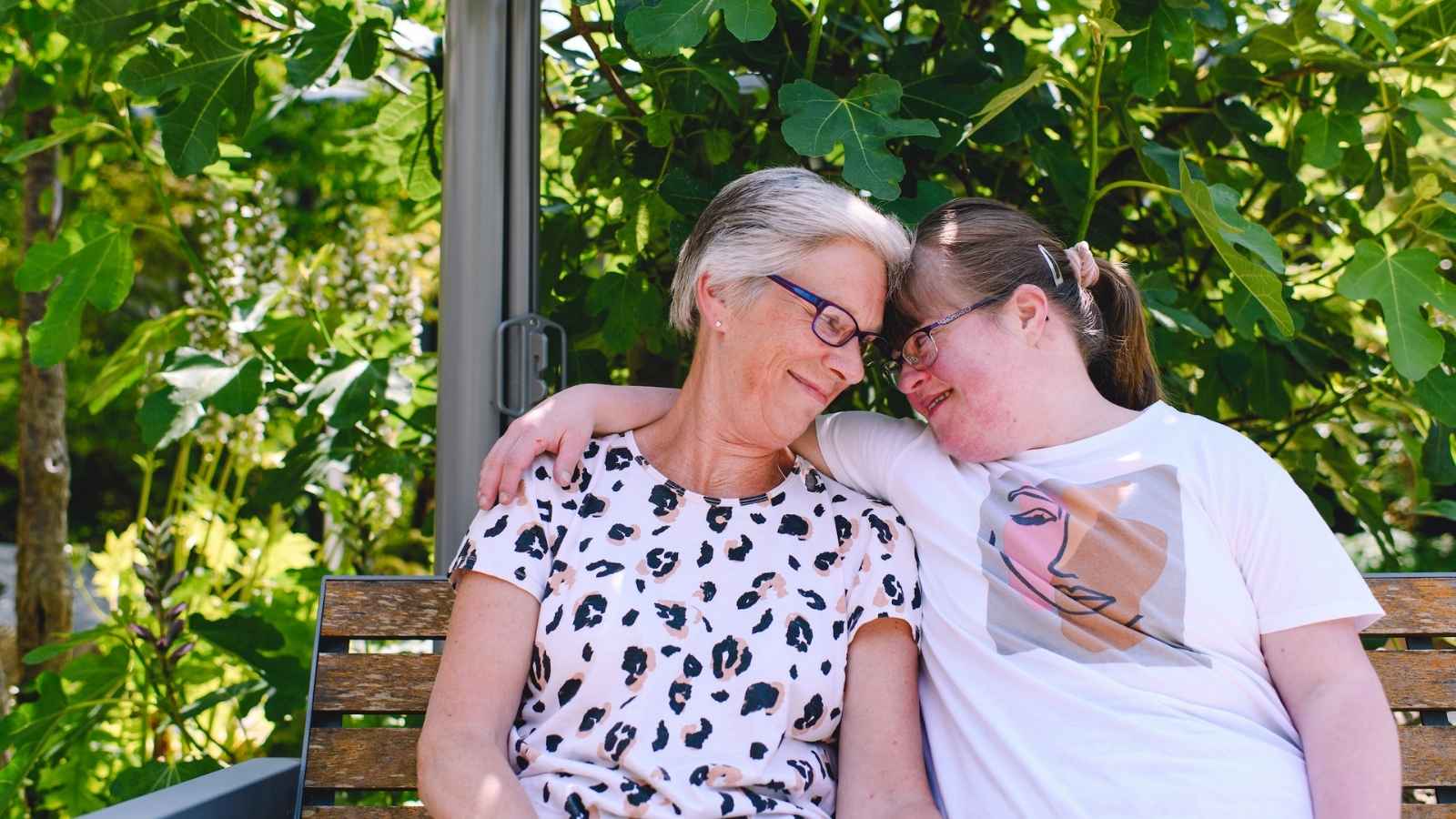 Two women sit on a bench in a green garden, one with their arm around the other, and they rest their foreheads together.