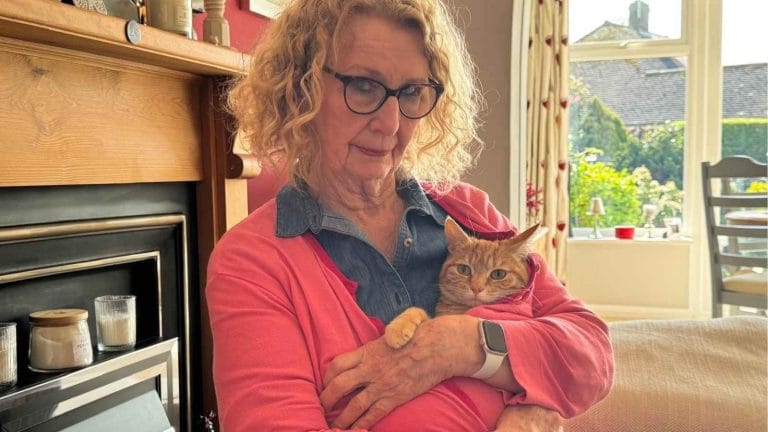 A woman stands in a pink cardigan holding a ginger cat who is wrapped also in the cardigan.