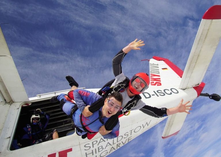 Two people in skydiving gear jumping out of a plane in tandem
