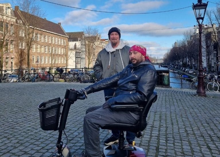 Dan and Carl on holiday by a canal in Amsterdam, Dan is in a mobility scooter
