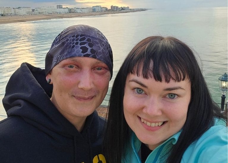 Carl and Marie on a pier with a calm sea behind them. They both smile at the camera