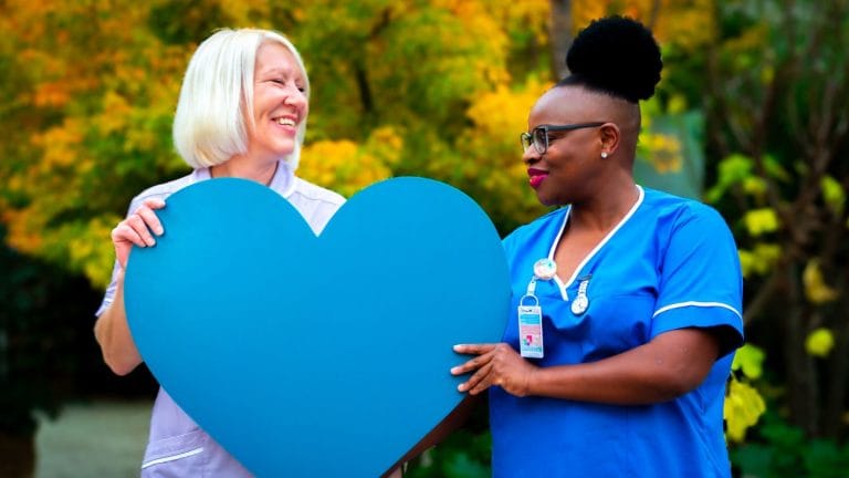 Two members of the care team at St Barnabas House outdoors and smiling while holding a big blue heart